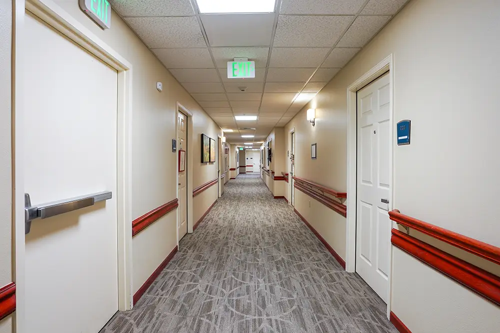 Long, empty hallway in a building with beige walls, red handrails, closed white doors, overhead lights, and framed art on the walls. Long, empty hallway in a building with beige walls, red handrails, closed white doors, overhead lights, and framed art on the walls.