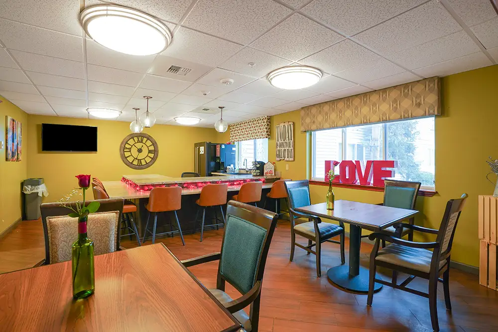 A brightly lit dining area with two tables, chairs, a counter with barstools, wall clock, window, and red “LOVE” letters on the windowsill. A brightly lit dining area with two tables, chairs, a counter with barstools, wall clock, window, and red “LOVE” letters on the windowsill.