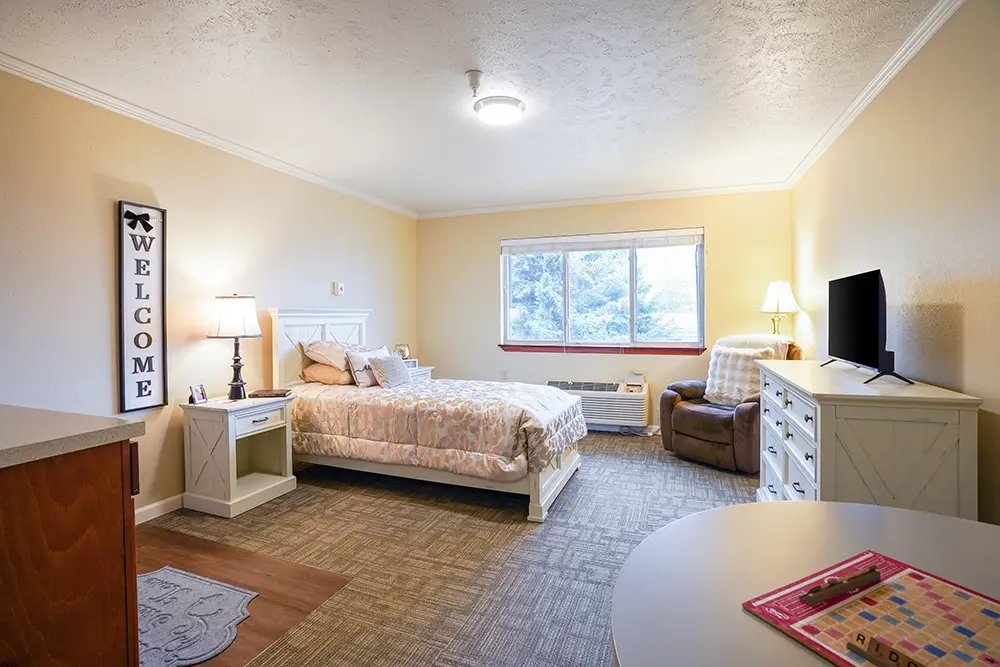 A neatly arranged bedroom with a single bed, dresser, armchair, TV, and a window. A “Welcome” sign and a board game on a round table are also visible. A neatly arranged bedroom with a single bed, dresser, armchair, TV, and a window. A "Welcome" sign and a board game on a round table are also visible.