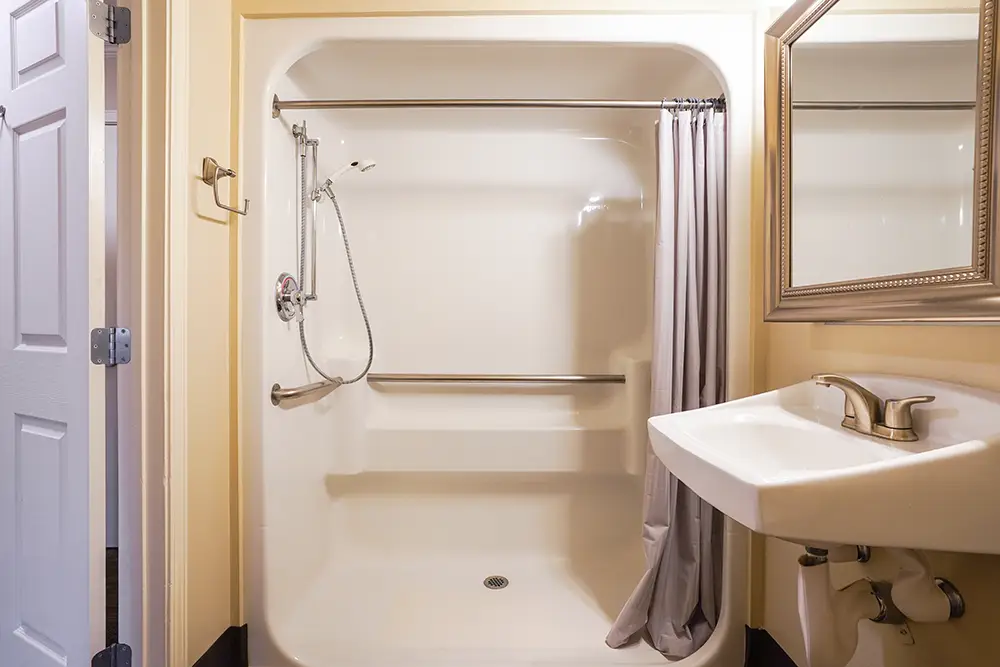 Accessible shower with grab bars, handheld showerhead, shower curtain, towel rack, wall-mounted sink, and a mirror in a bathroom with light-colored walls. Accessible shower with grab bars, handheld showerhead, shower curtain, towel rack, wall-mounted sink, and a mirror in a bathroom with light-colored walls.