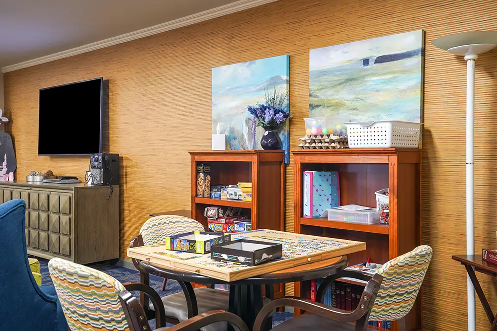 A game table with board games and puzzles, surrounded by four chairs, sits in a cozy room with bookshelves, abstract paintings, and a TV mounted on a tan wall. A game table with board games and puzzles, surrounded by four chairs, sits in a cozy room with bookshelves, abstract paintings, and a TV mounted on a tan wall.