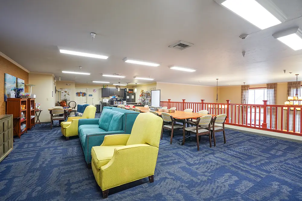 A spacious common room with blue carpet, yellow armchairs, a teal sofa, a dining table with chairs, shelves, and bright ceiling lights. The area overlooks a lower level with red railings. A spacious common room with blue carpet, yellow armchairs, a teal sofa, a dining table with chairs, shelves, and bright ceiling lights. The area overlooks a lower level with red railings.