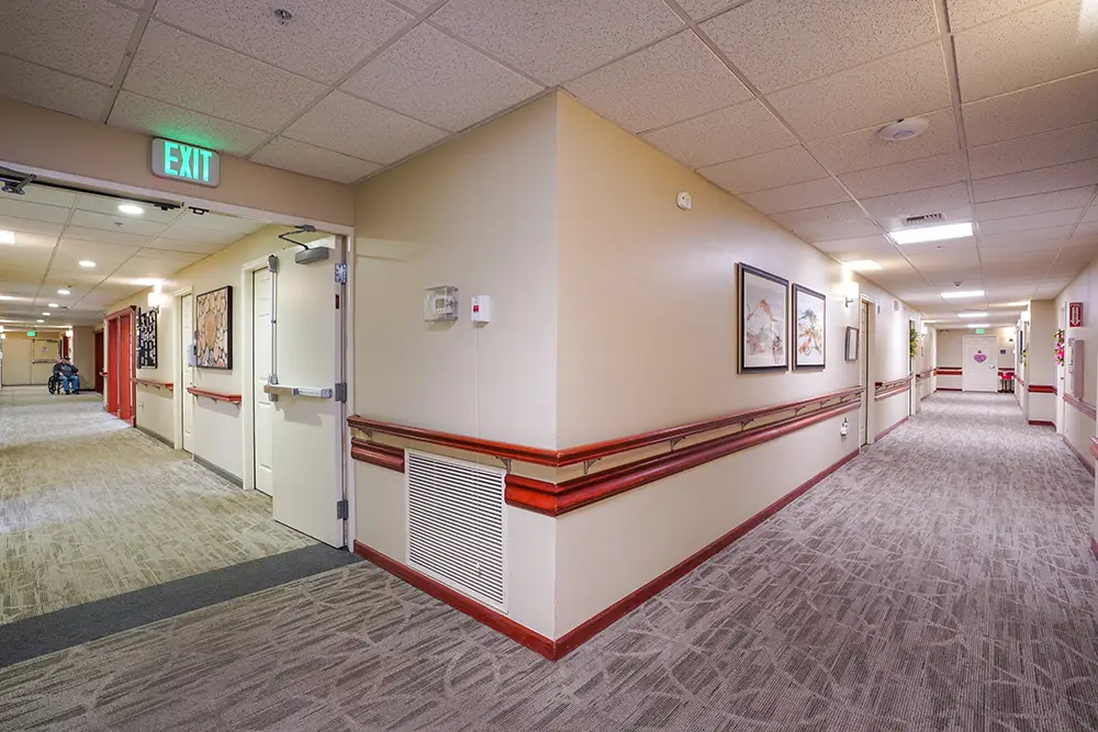 A well-lit hallway in a building with beige walls, carpeted floor, framed artwork, handrails, exit sign, and doors along the corridor. A well-lit hallway in a building with beige walls, carpeted floor, framed artwork, handrails, exit sign, and doors along the corridor.