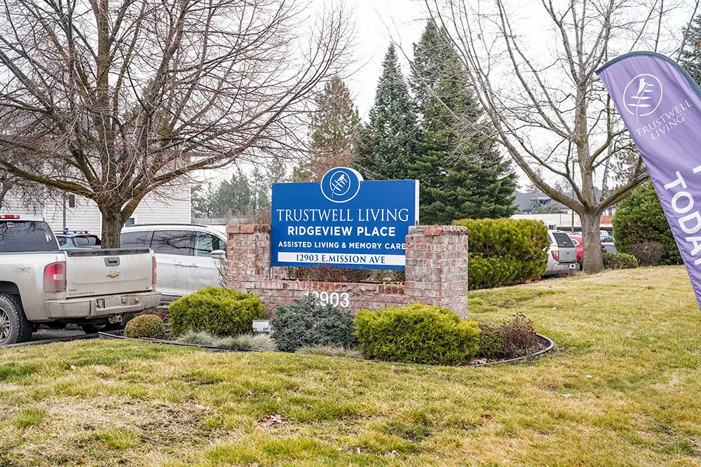 A brick sign for Trustwell Living Ridgeview Place Assisted Living & Memory Care, with contact info, stands on a grassy lawn near parked vehicles and trees. A brick sign for Trustwell Living Ridgeview Place Assisted Living & Memory Care, with contact info, stands on a grassy lawn near parked vehicles and trees.