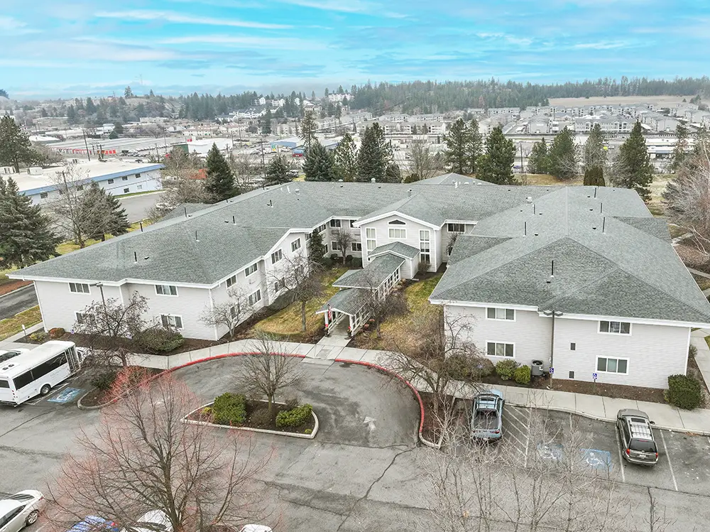 Aerial view of a large, white, two-story building with a gray roof, surrounded by trees, parked cars, and a parking lot on a cloudy day. Aerial view of a large, white, two-story building with a gray roof, surrounded by trees, parked cars, and a parking lot on a cloudy day.