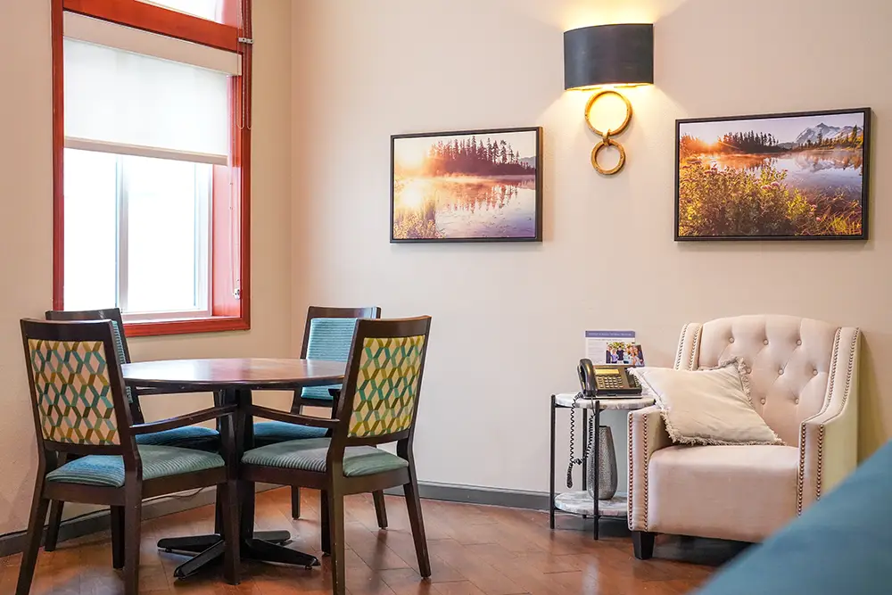 A small dining area with a round table and chairs near a window, next to an armchair, side table, and two landscape photos on the wall. A small dining area with a round table and chairs near a window, next to an armchair, side table, and two landscape photos on the wall.