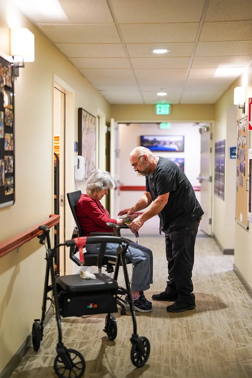 A caregiver assists an elderly person seated in a wheelchair in a well-lit hallway; a walker is nearby. A caregiver assists an elderly person seated in a wheelchair in a well-lit hallway; a walker is nearby.