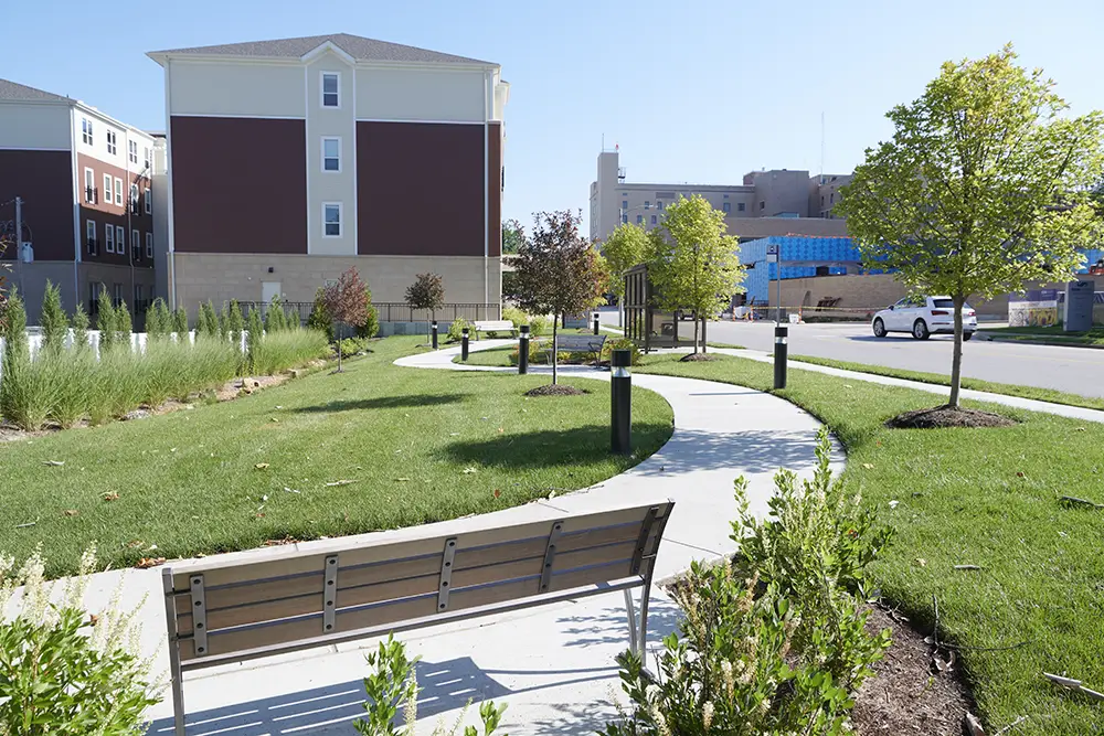 Trustwell Living at Richmond Heights Exterior 3 A curved sidewalk runs through a small grassy park with trees, shrubs, and a bench, surrounded by multi-story buildings on a sunny day.