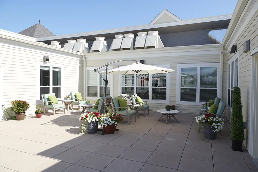Trustwell Living at Richmond Heights Exterior 4 A sunny patio with potted flowers, green-cushioned chairs, small tables, and a large white umbrella, surrounded by cream-colored building walls and windows.