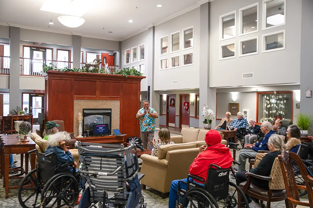 A group of seniors, some in wheelchairs, sit in a lounge area watching a man speaking in front of a fireplace in a retirement home. A group of seniors, some in wheelchairs, sit in a lounge area watching a man speaking in front of a fireplace in a retirement home.