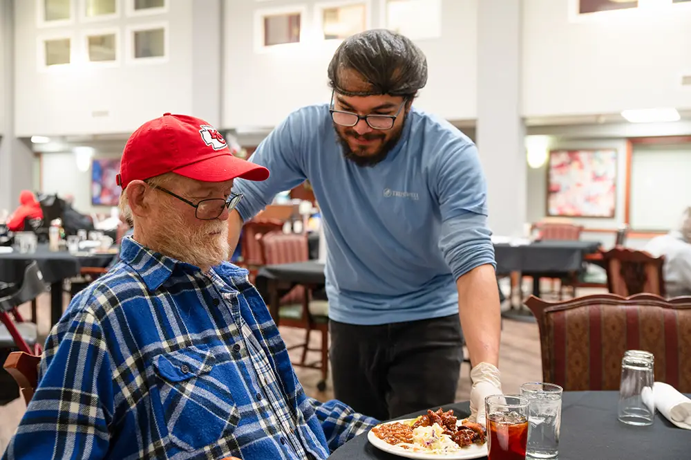 A man in a red hat sits at a table while another man in glasses and gloves serves him a plate of food in a dining hall. A man in a red hat sits at a table while another man in glasses and gloves serves him a plate of food in a dining hall.