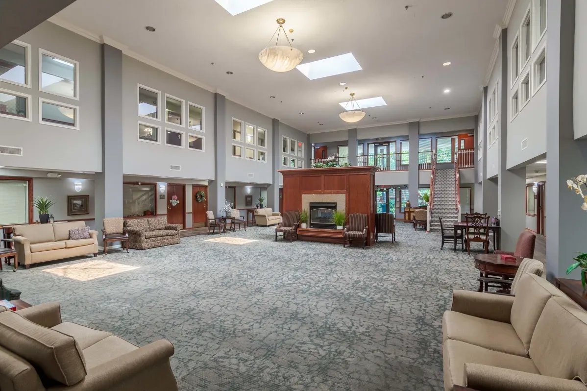Spacious, well-lit lobby area at Trustwell Senior Living with sofas, armchairs, a central fireplace, carpeted floor, skylights, and a staircase leading to an upper level.