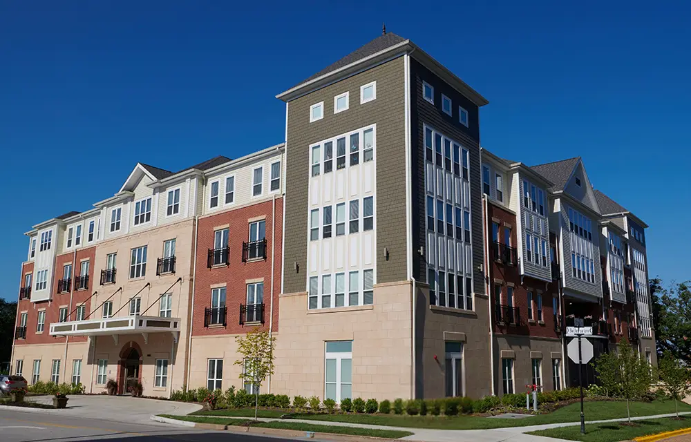Trustwelll Living at Richmond Heights Outdoor Shot A modern four-story apartment building with brick and siding exterior, large windows, and a small landscaped area in front under a clear blue sky.