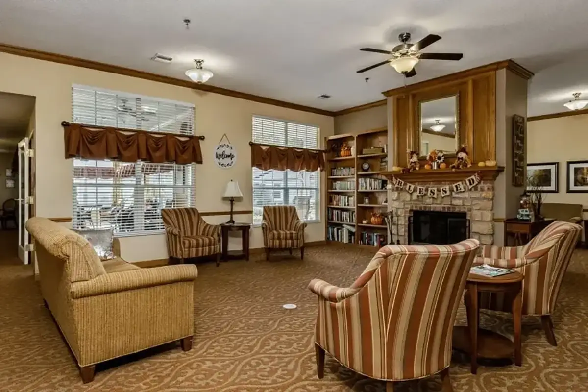 A cozy living room at Trustwell Senior Living features armchairs, a sofa, a fireplace, bookshelves, and large windows. Ceiling fans and neutral colors create a warm, inviting atmosphere perfect for relaxation.