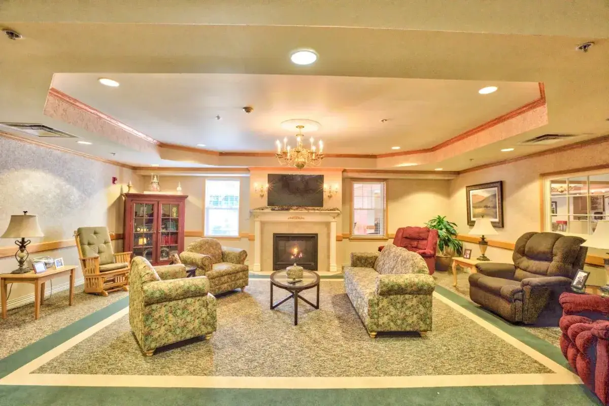 A cozy Trustwell Senior Living lounge with armchairs, sofas, and a fireplace, featuring a chandelier, TV above the mantle, and windows letting in natural light.
