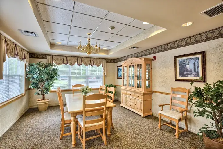 A dining room at Trustwell Senior Living features a wooden table, six chairs, a china cabinet, potted plants, and large windows with valances that let in natural light. A dining room at Trustwell Senior Living features a wooden table, six chairs, a china cabinet, potted plants, and large windows with valances that let in natural light.