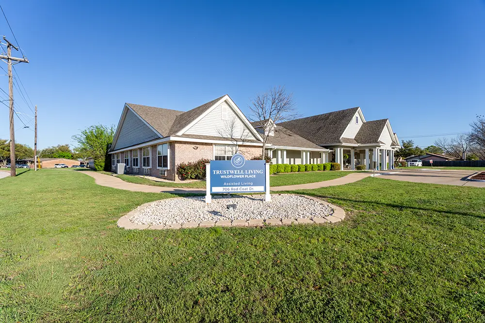 A single-story assisted living facility with a gabled roof, white exterior, and a sign in front reading “Trustwell Living.” The building is surrounded by grass and a clear blue sky. A single-story assisted living facility with a gabled roof, white exterior, and a sign in front reading "Trustwell Living." The building is surrounded by grass and a clear blue sky.