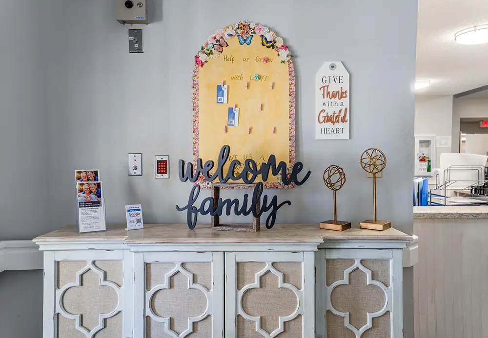 A white cabinet with a “welcome family” sign, gratitude wall, brochures, and gold decorative items in a home or facility entryway. A white cabinet with a “welcome family” sign, gratitude wall, brochures, and gold decorative items in a home or facility entryway.