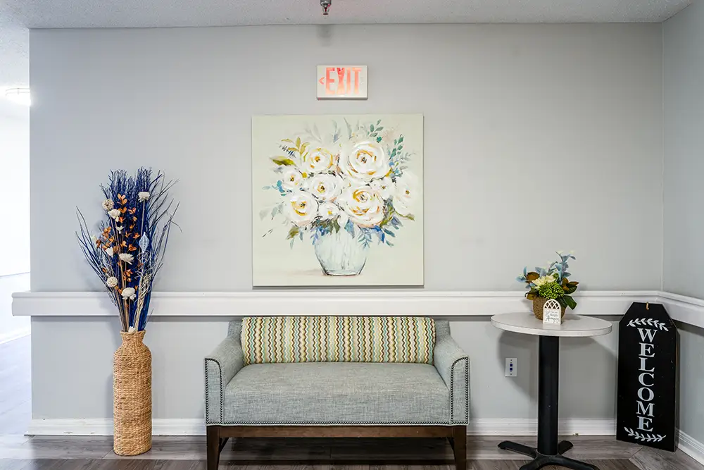 A small bench sits below a painting of white flowers; a tall vase, side table with flowers, and a black “Welcome” sign are nearby under an exit sign. A small bench sits below a painting of white flowers; a tall vase, side table with flowers, and a black "Welcome" sign are nearby under an exit sign.