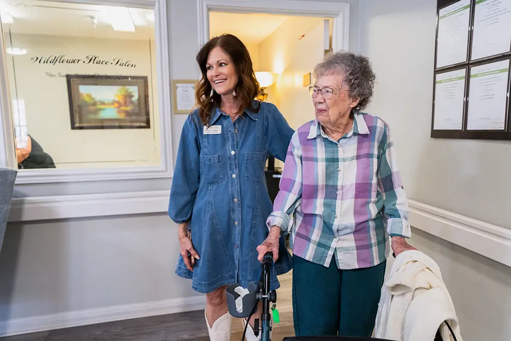 A younger woman stands beside an older woman using a walker in a brightly lit room. Both are smiling and looking in the same direction. A younger woman stands beside an older woman using a walker in a brightly lit room. Both are smiling and looking in the same direction.