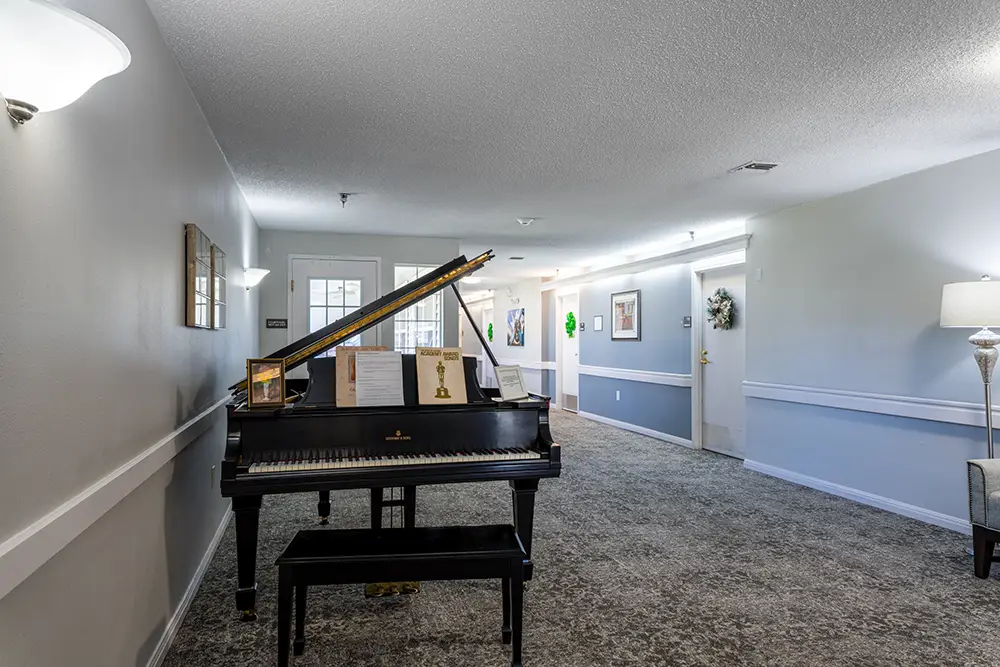 Black grand piano with sheet music on a stand in a carpeted hallway with gray walls, wall lamps, and closed doors visible in the background. Black grand piano with sheet music on a stand in a carpeted hallway with gray walls, wall lamps, and closed doors visible in the background.
