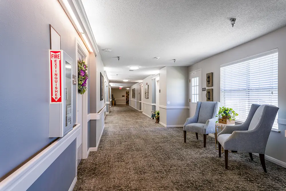 A carpeted hallway with light gray walls, two armchairs and a table by a window, and a fire extinguisher sign on the left wall. A carpeted hallway with light gray walls, two armchairs and a table by a window, and a fire extinguisher sign on the left wall.