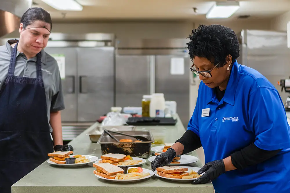 Two cafeteria workers assemble plates of food on a counter in a commercial kitchen, with sandwiches and sides arranged on several plates. Two cafeteria workers assemble plates of food on a counter in a commercial kitchen, with sandwiches and sides arranged on several plates.