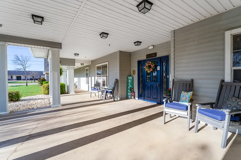 Covered front porch with gray siding, blue door, rocking chairs with cushions, a “Welcome” sign, and decorative wreath. Grass lawn and sidewalk visible in the background. Covered front porch with gray siding, blue door, rocking chairs with cushions, a "Welcome" sign, and decorative wreath. Grass lawn and sidewalk visible in the background.