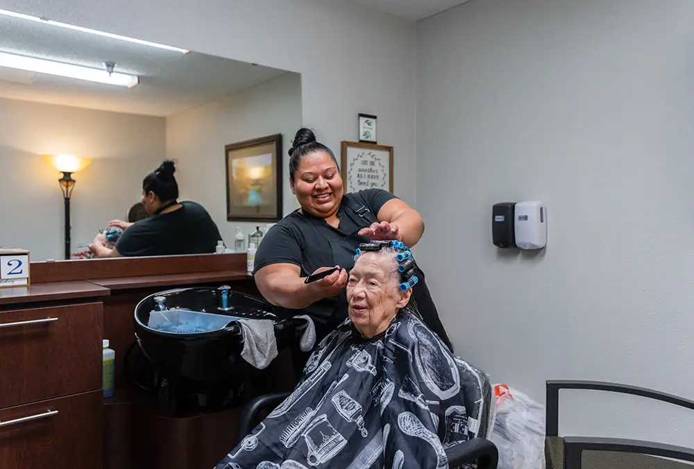 A hairdresser applies hair dye to an elderly person with curlers in their hair, seated in a salon chair. Both are in a well-lit salon with a mirror and sink in the background. A hairdresser applies hair dye to an elderly person with curlers in their hair, seated in a salon chair. Both are in a well-lit salon with a mirror and sink in the background.