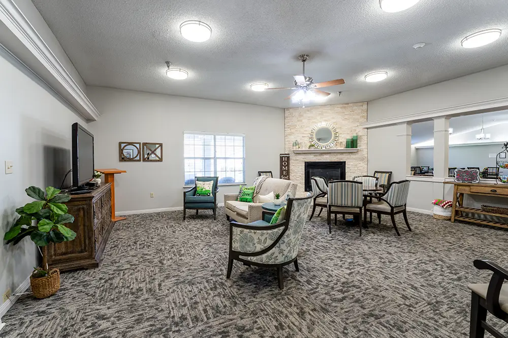 A living room with a TV, chairs, sofa, table, fireplace, and several decorations. Large window and ceiling lights provide bright illumination. A living room with a TV, chairs, sofa, table, fireplace, and several decorations. Large window and ceiling lights provide bright illumination.