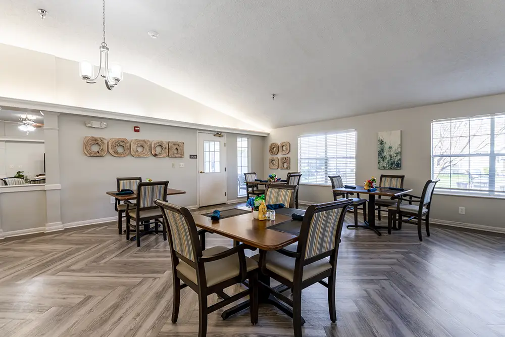 A dining area with four square tables, each with four chairs, neutral walls, large windows, wood-patterned floor, and wall art decorations. A dining area with four square tables, each with four chairs, neutral walls, large windows, wood-patterned floor, and wall art decorations.