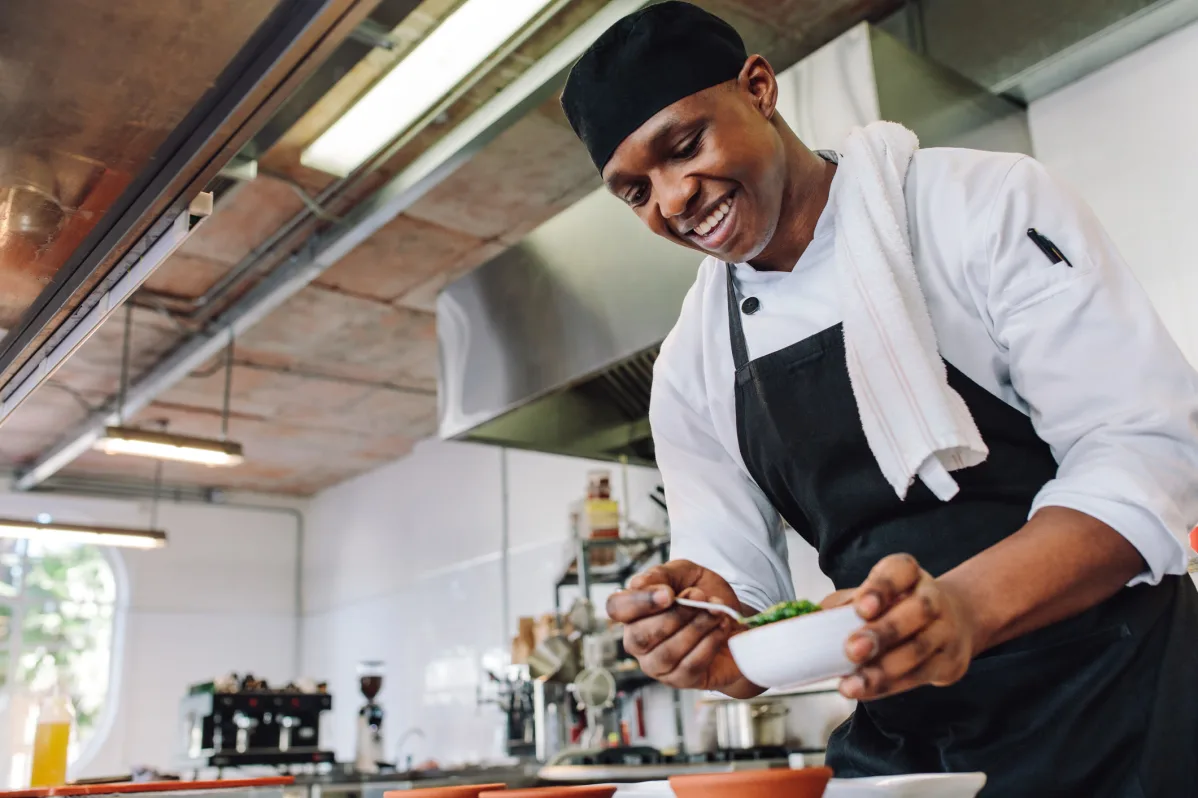 A chef in a white uniform and black apron smiles while garnishing a dish in the Trustwell Senior Living commercial kitchen. A chef in a white uniform and black apron smiles while garnishing a dish in the Trustwell Senior Living commercial kitchen.