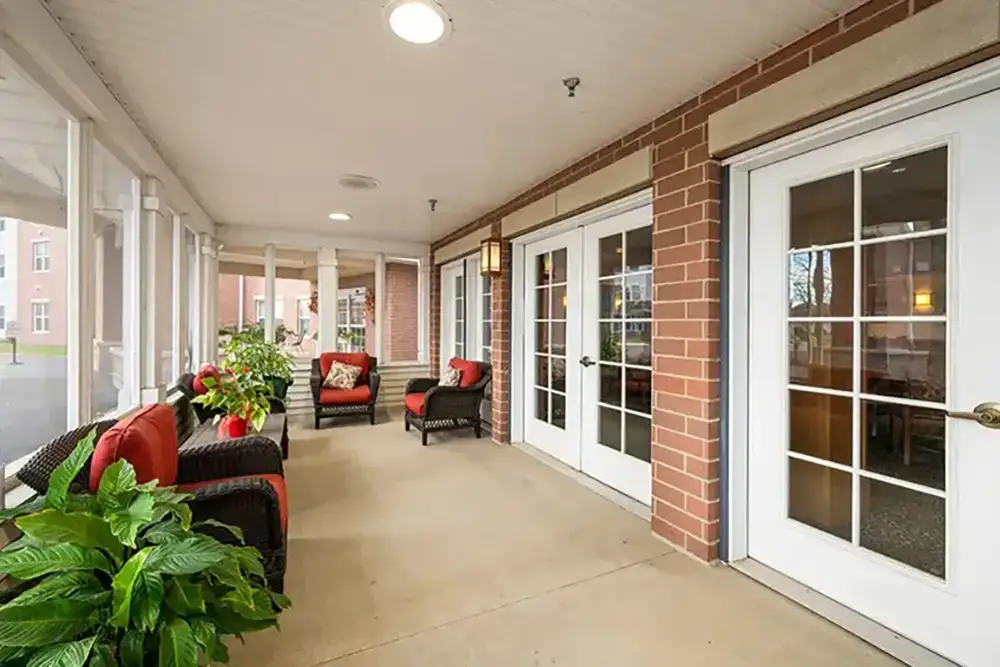 Covered patio with wicker chairs and red cushions, several potted plants, and large glass doors along a brick wall create a welcoming outdoor space at Trustwell Senior Living.