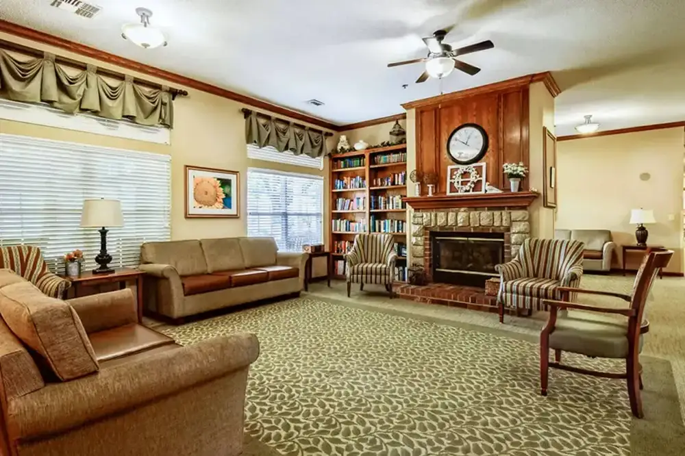A welcoming living room at Trustwell Senior Living features sofas, armchairs, a fireplace, built-in bookshelves, a large clock, and patterned carpet, warmly lit by ceiling fans and table lamps.