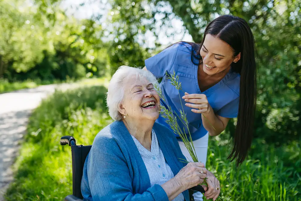 A smiling elderly woman in a wheelchair holds wildflowers while a younger woman in scrubs from Trustwell Senior Living leans over her; they are outdoors on a sunny day. A smiling elderly woman in a wheelchair holds wildflowers while a younger woman in scrubs from Trustwell Senior Living leans over her; they are outdoors on a sunny day.