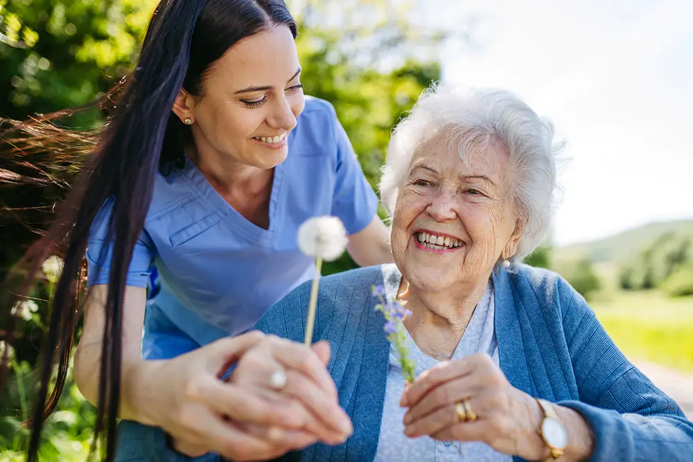 A young woman assists an elderly woman holding flowers outdoors at Trustwell Senior Living; both are smiling in a sunny, green setting.