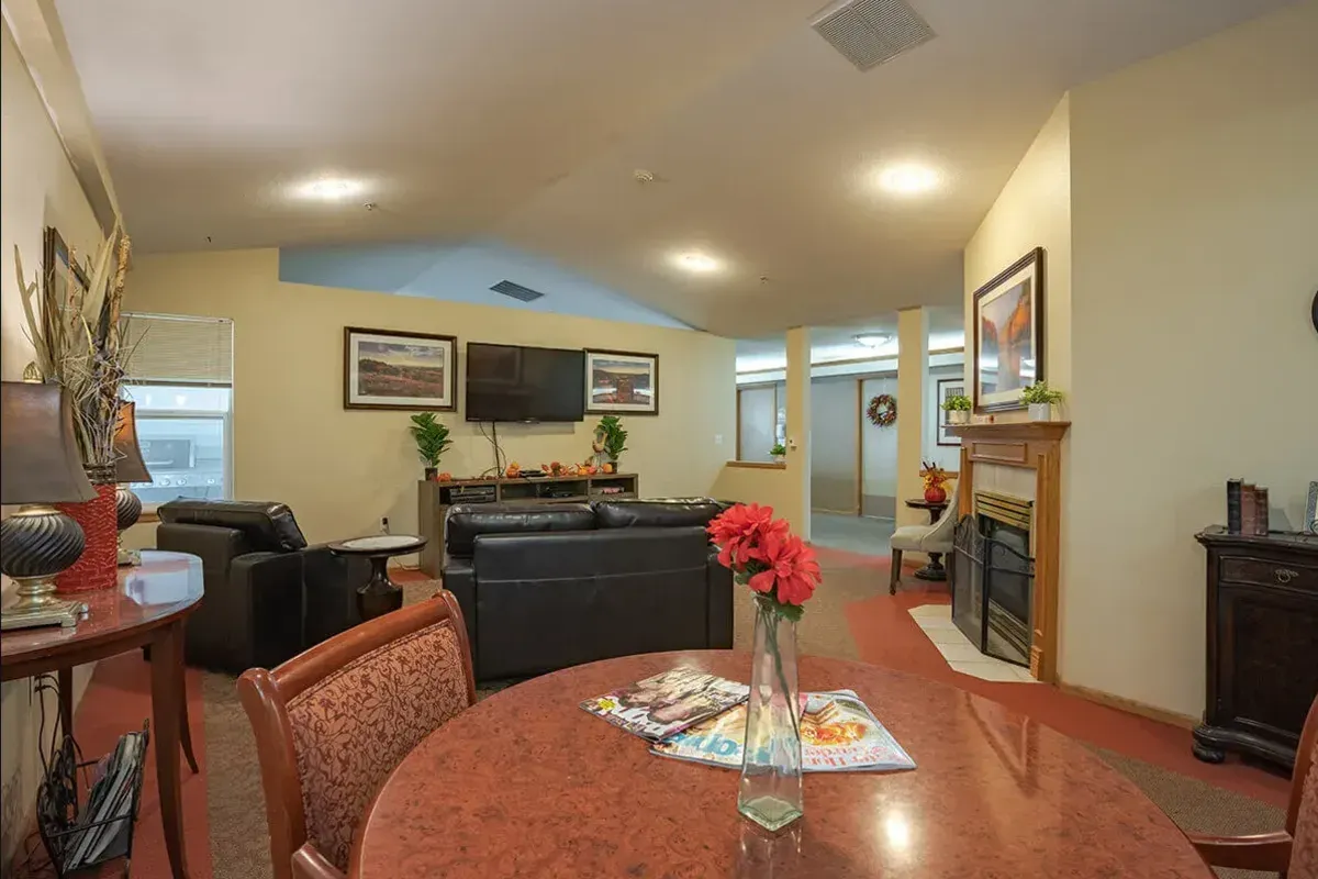 A cozy communal living area at Trustwell Senior Living features a round table, leather sofas, a fireplace, TV, and charming touches like flowers and framed pictures.