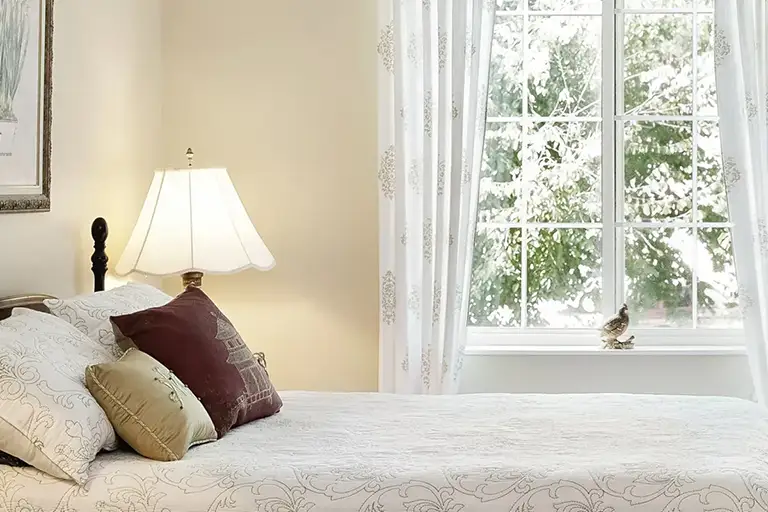 Bedroom at Trustwell Senior Living with a neatly made bed, several pillows, a bedside lamp, and a window with white curtains revealing snow-covered trees outside. Bedroom at Trustwell Senior Living with a neatly made bed, several pillows, a bedside lamp, and a window with white curtains revealing snow-covered trees outside.