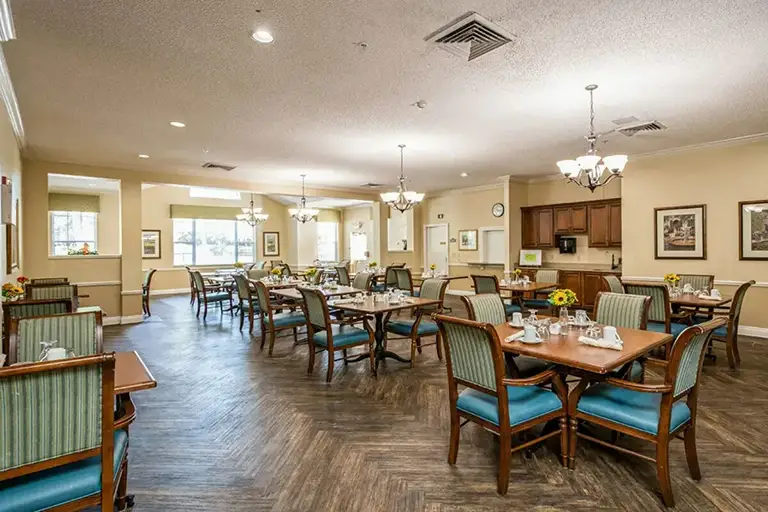 A well-lit dining room at Trustwell Senior Living features wooden tables and chairs, elegant place settings, chandeliers, and framed artwork on cream-colored walls. A well-lit dining room at Trustwell Senior Living features wooden tables and chairs, elegant place settings, chandeliers, and framed artwork on cream-colored walls.