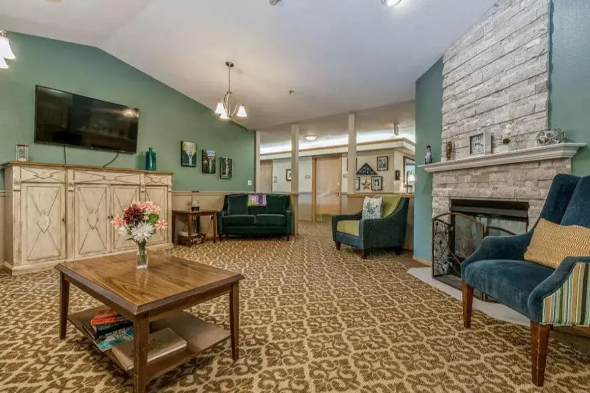 A living room at Trustwell Senior Living features a patterned carpet, a coffee table with flowers, two armchairs, a green sofa, a cabinet with a TV, and a cozy fireplace with a stone surround.