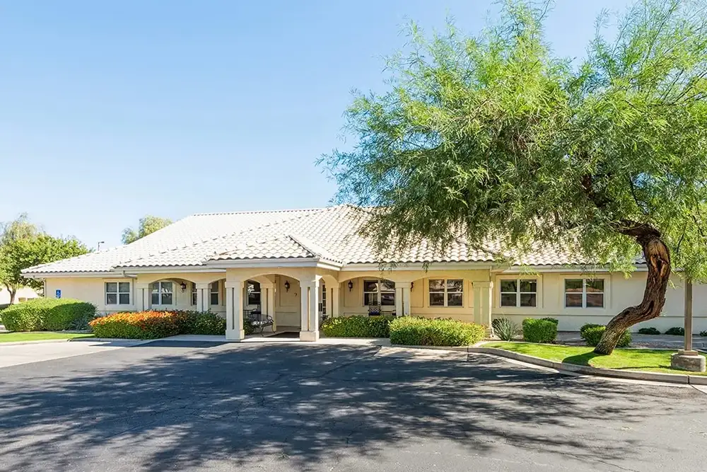 Single-story Trustwell Senior Living building with a tiled roof, multiple windows, and a covered entrance, surrounded by landscaping and a paved parking area under a clear sky.