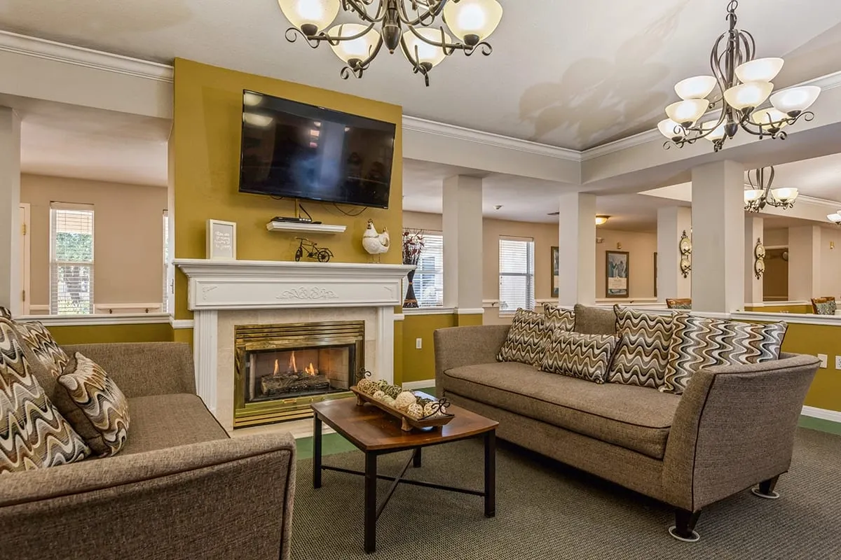 A Trustwell Senior Living living room with two brown sofas, a wooden coffee table, a lit fireplace, wall-mounted TV, and multiple chandeliers, set against mustard yellow and white walls.
