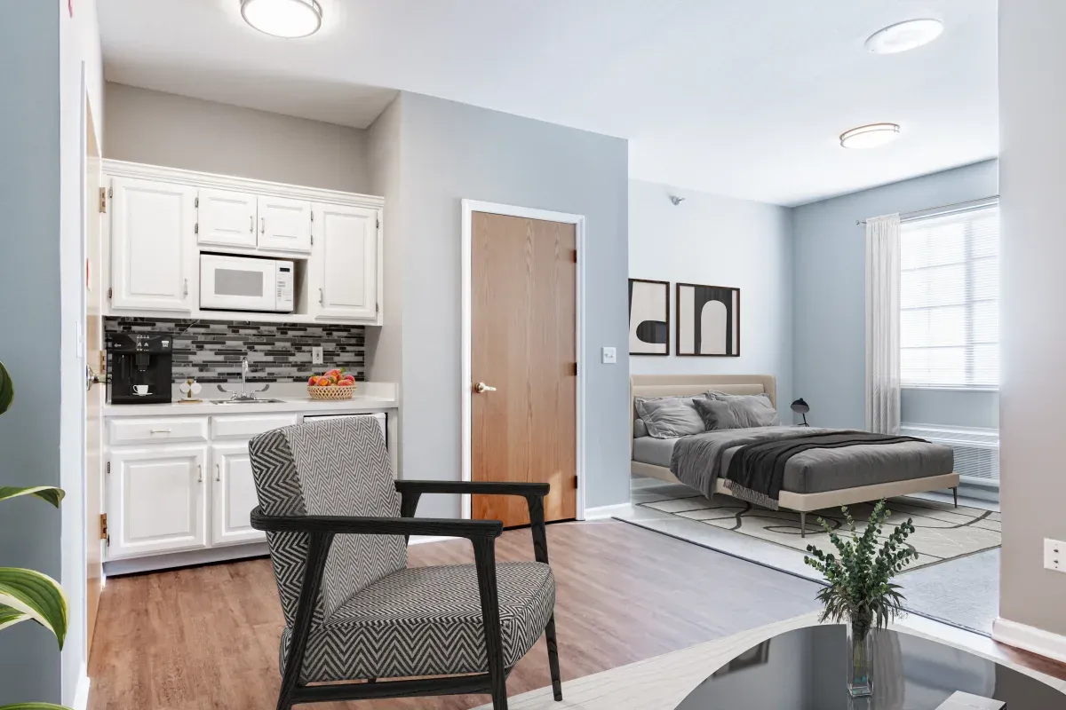 A modern studio apartment at Trustwell Senior Living with a small kitchen, a bed, a chair, and a table; neutral colors and natural light from a large window.
