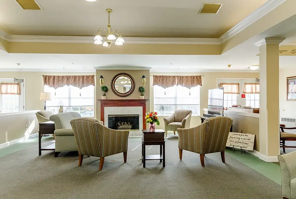 A well-lit communal living room at Trustwell Senior Living features armchairs, a fireplace with a mirror above it, side tables, and large windows letting in natural light.
