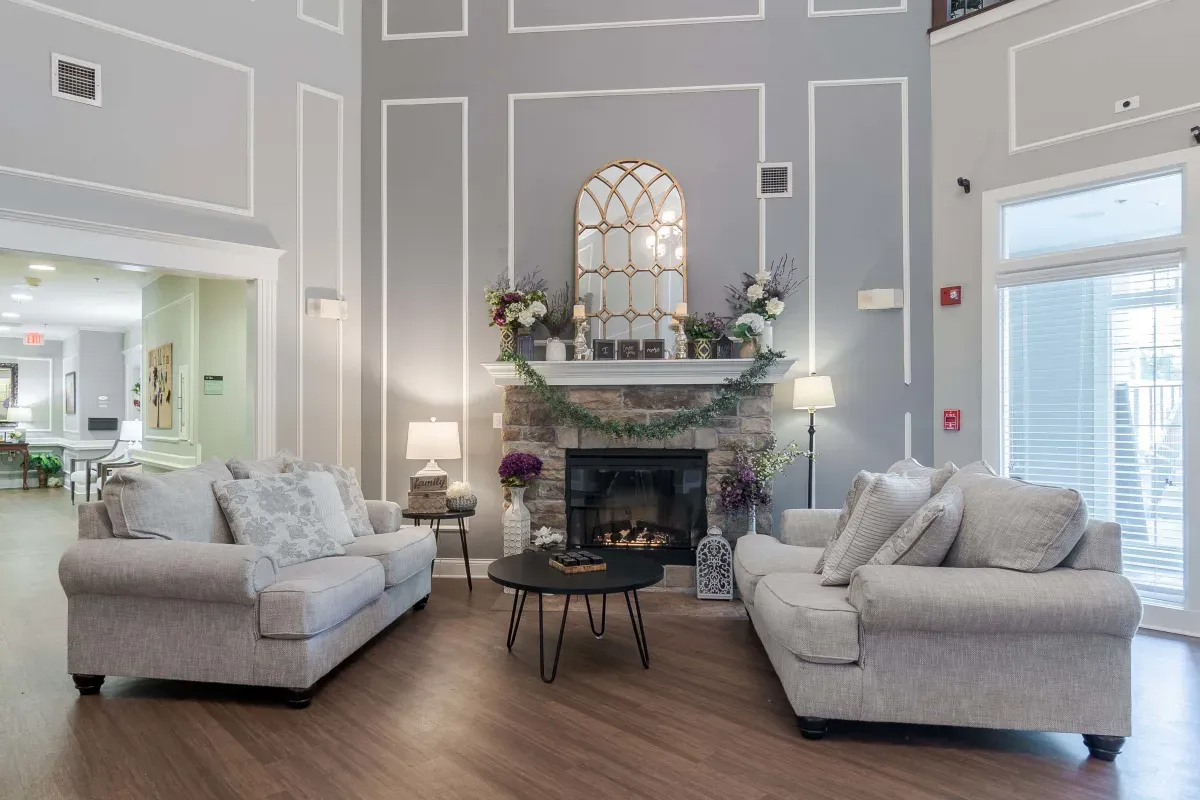 A living room at Trustwell Senior Living features two gray sofas facing a fireplace, a circular black coffee table, and a large arched mirror above the mantel adorned with greenery and flowers.