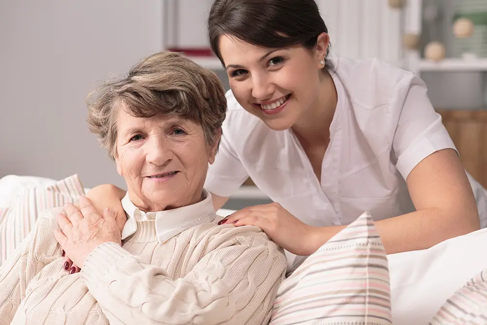 An older woman sits on a couch while a younger woman in a white uniform stands behind her, both smiling at the camera. An older woman sits on a couch while a younger woman in a white uniform stands behind her, both smiling at the camera. An older woman sits on a couch while a younger woman in a white uniform stands behind her, both smiling at the camera.