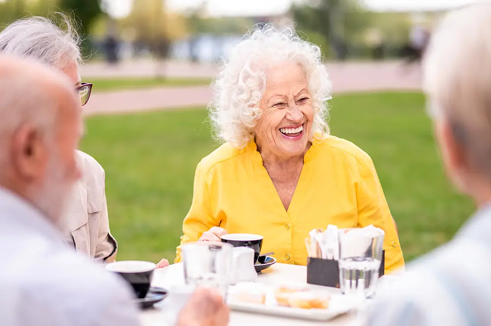 An elderly woman in a bright yellow shirt smiles while sitting at an outdoor table with three other people, drinking coffee in a park setting.