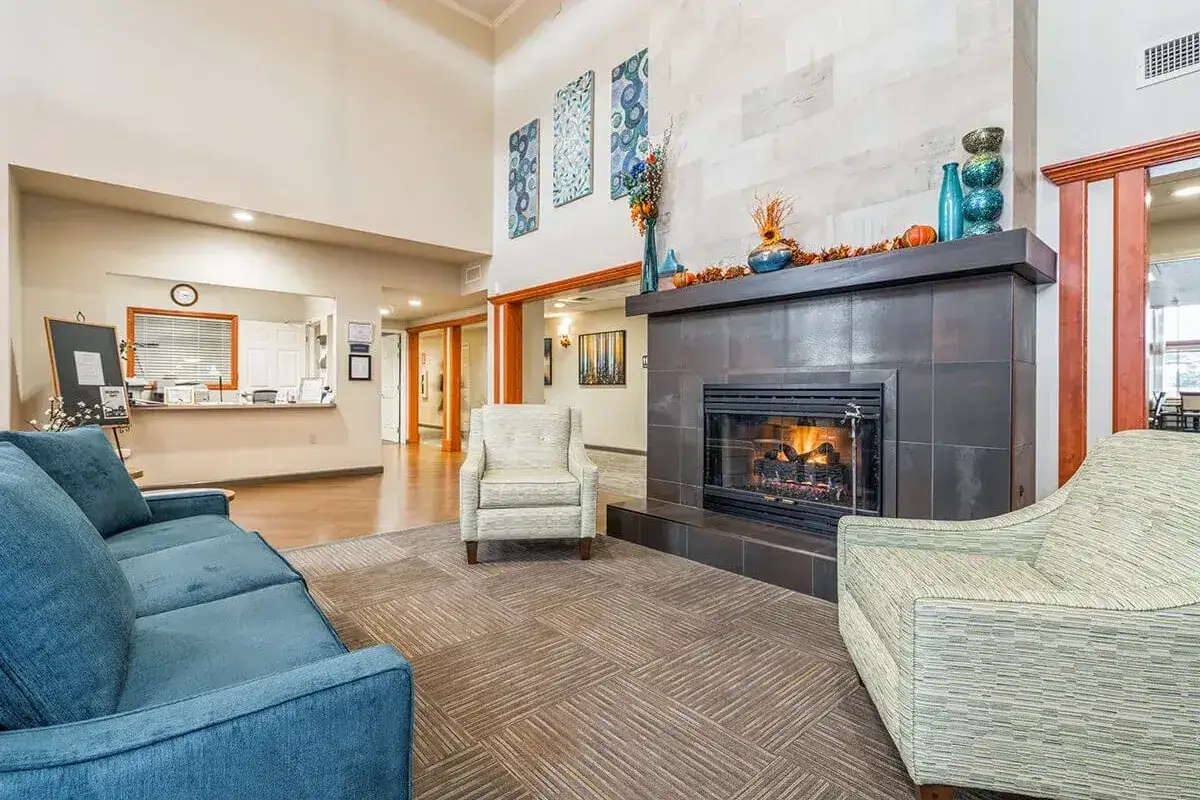 A modern Trustwell Senior Living lobby featuring a fireplace, blue and beige armchairs, a blue sofa, autumn décor on the mantel, and wall art above the fireplace. A welcoming reception desk is visible in the background.