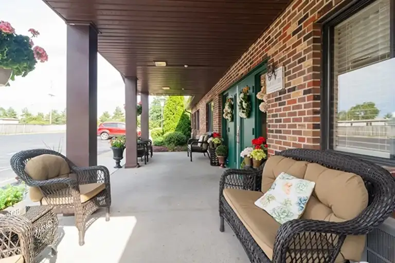 A covered brick porch at Trustwell Senior Living features cushioned wicker chairs, potted plants, and decorative wreaths on the doors, overlooking a parking lot and greenery. A covered brick porch at Trustwell Senior Living features cushioned wicker chairs, potted plants, and decorative wreaths on the doors, overlooking a parking lot and greenery.