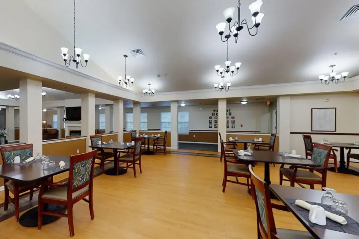 A spacious dining area at Trustwell Senior Living features wooden floors, several tables and chairs, place settings, chandeliers, and neutral-colored walls. The room is empty and well-lit.